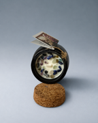 An open black cauldron-shaped candle with visible wax and colorful glass stones, resting on a cork base.