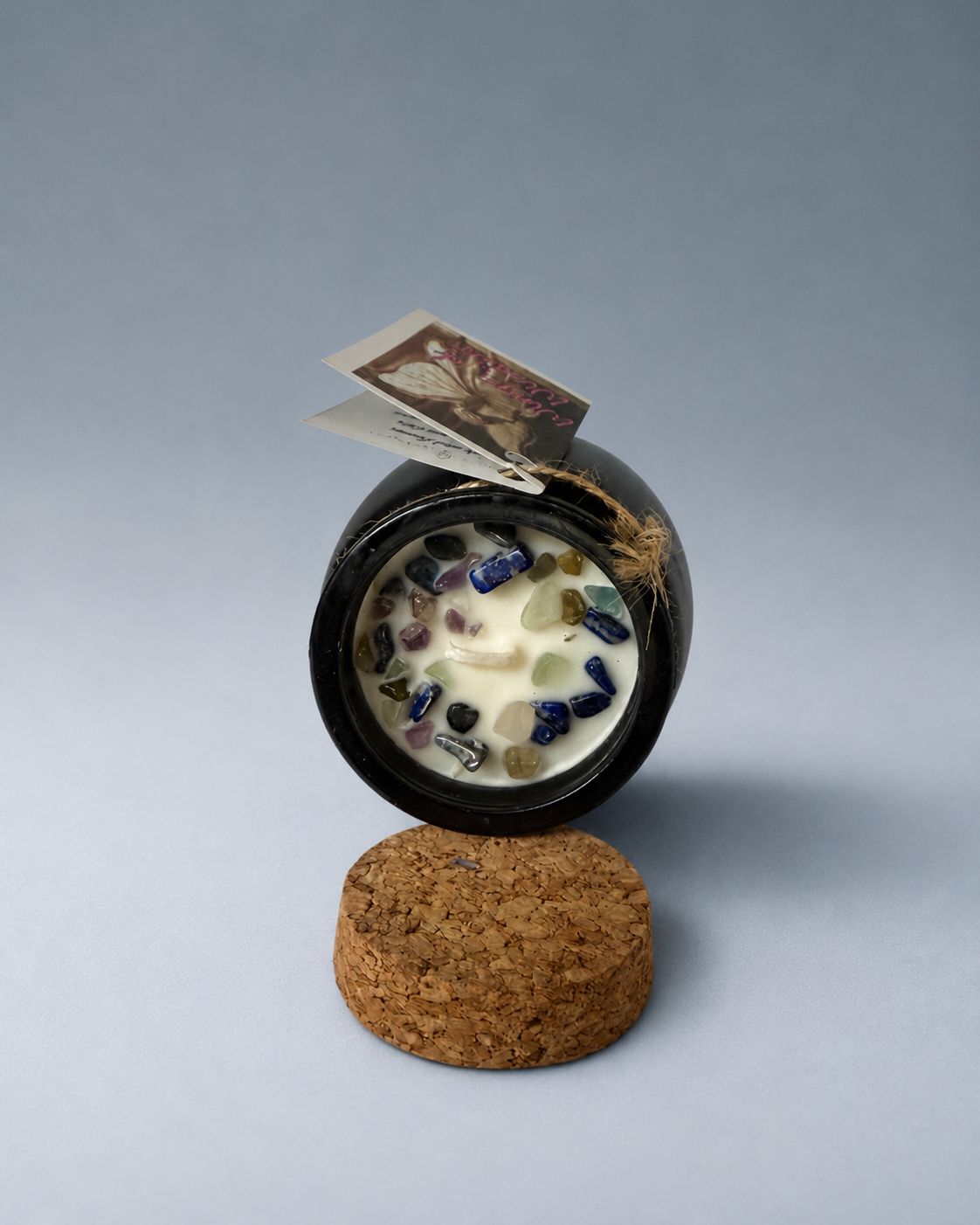 An open black cauldron-shaped candle with visible wax and colorful glass stones, resting on a cork base.