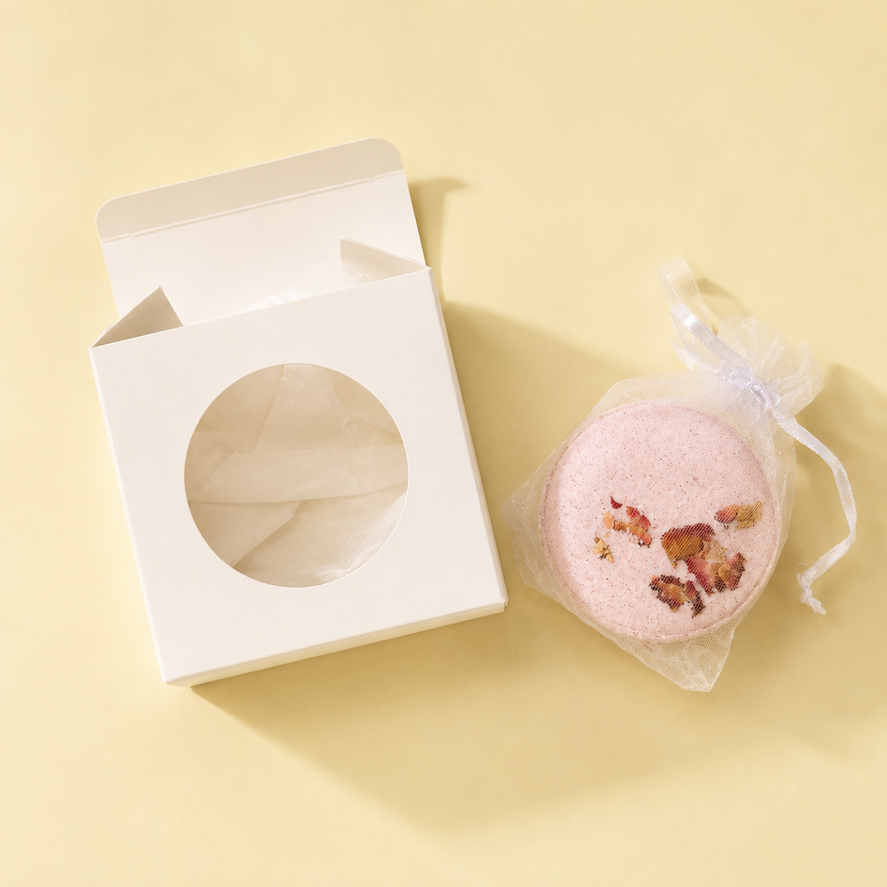 An open box beside a Joy bath bomb in a mesh drawstring bag on a yellow background.