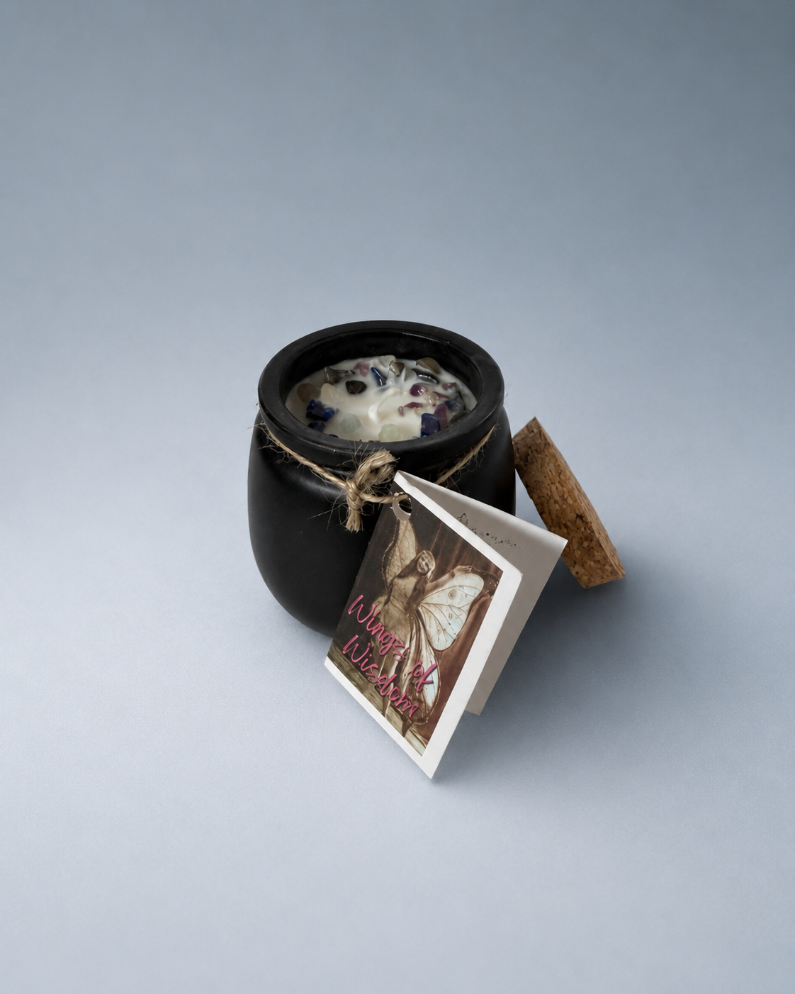An open black cauldron-shaped candle with a cork lid beside it and a 'Wings of Wisdom' tag.