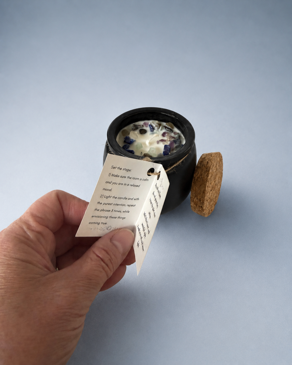 A hand holding a printed intention tag next to an open black cauldron-shaped candle with a cork lid.