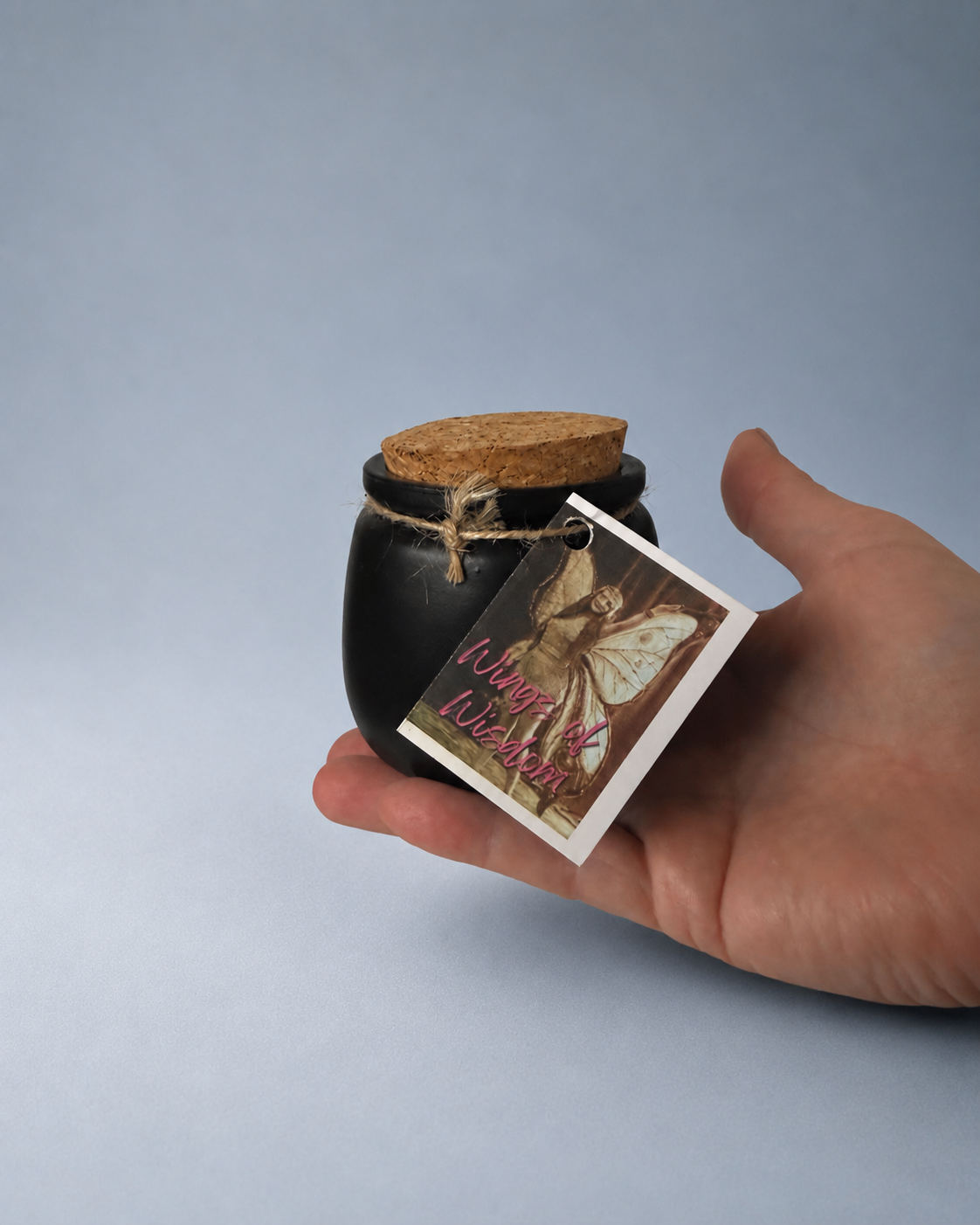 A hand holding a small black jar with a cork lid and a 'Wings of Wisdom' tag.