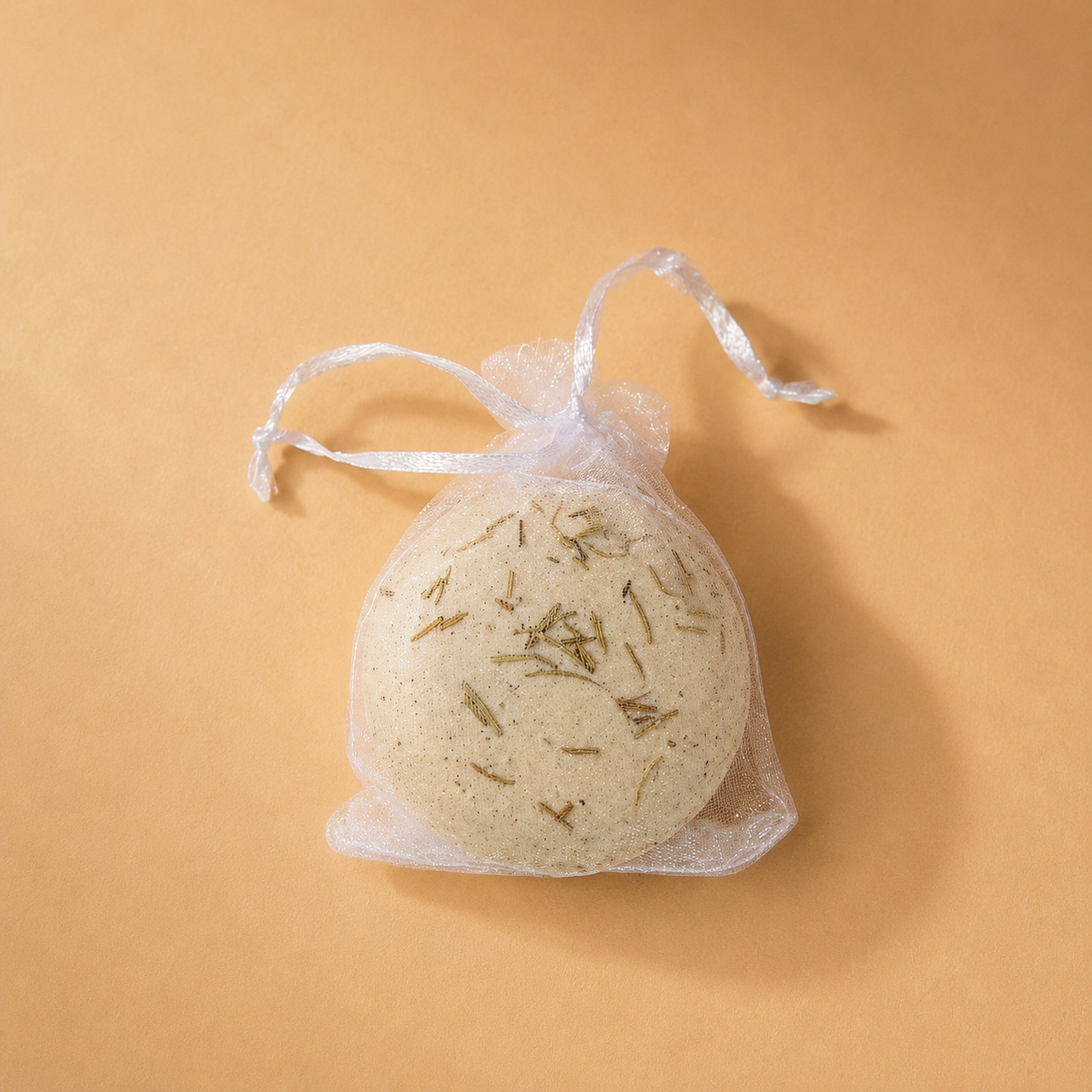 A beige Ground bath bomb with dried herbs in a mesh drawstring bag.