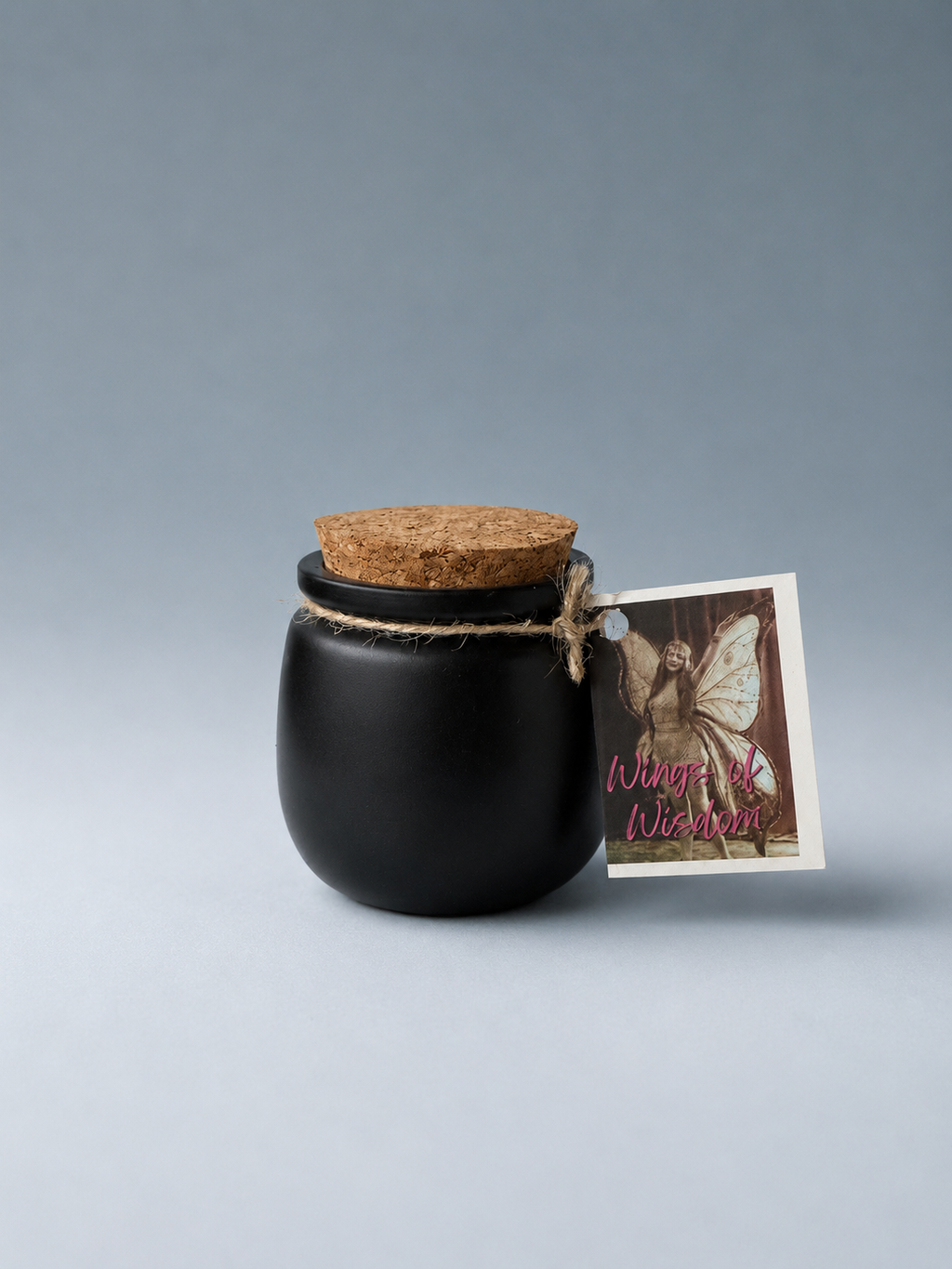 A black jar with a cork lid and a 'Wings of Wisdom' tag, on a neutral gray background.