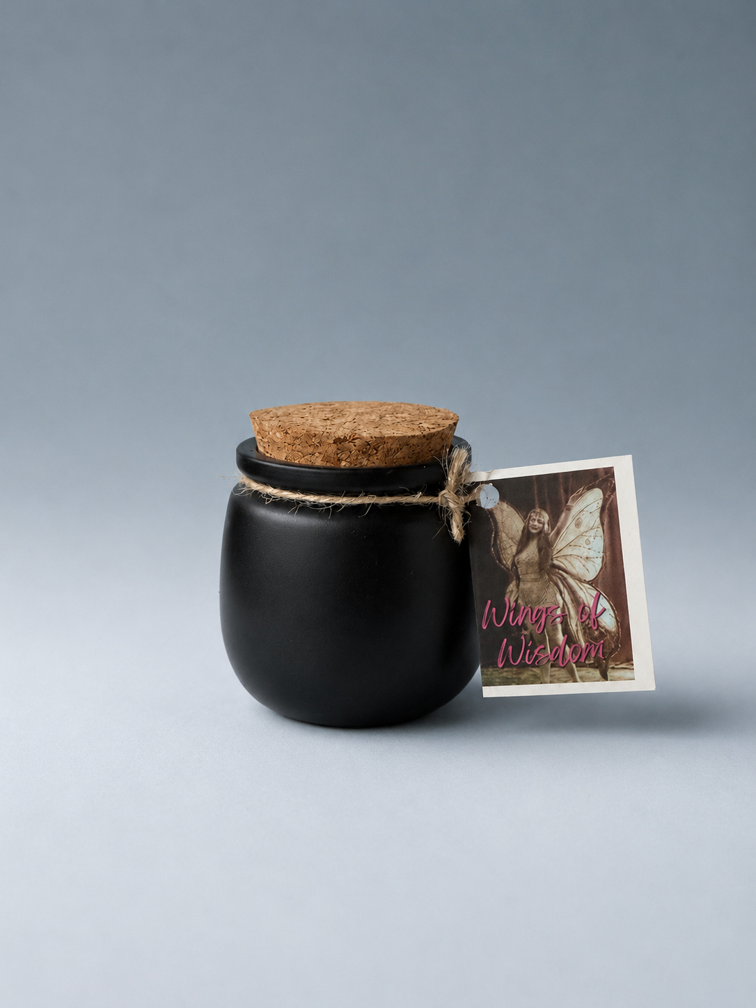 A black jar with a cork lid and a 'Wings of Wisdom' tag, on a neutral gray background.