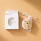 An open box beside a beige Ground bath bomb in a mesh drawstring bag.