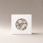 White box with a circular window showing dried botanical pieces inside, on a warm gray background.