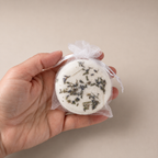 Hand holding a round Unwind bath bomb with visible lavender botanicals, wrapped in mesh and tied with ribbon.