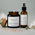 A lit Himalayan Orchid candle and room spray bottle, styled with orchid flowers and pink salt.
